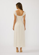 [Color: Natural] A back facing image of a brunette model wearing a beige linen blend maxi dress with short sleeves, a square neckline with tie detail, and a flowy skirt with a side slit. 