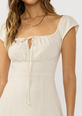 [Color: Natural] A detail image of a brunette model wearing a beige linen blend maxi dress with short sleeves, a square neckline with tie detail, and a flowy skirt with a side slit. 