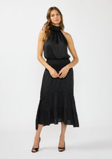 [Color: Black] A full body front facing image of a brunette model wearing a black holiday halter maxi dress with a sleeveless design, smocked drop waist, and tiered skirt. This flowy satin dress is perfect for any special occasion and holiday events. 