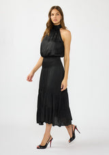 [Color: Black] A side facing image of a brunette model wearing a black holiday halter maxi dress with a sleeveless design, smocked drop waist, and tiered skirt. This flowy satin dress is perfect for any special occasion and holiday events. 