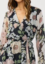 [Color: Black/Blush] A detail image of a brunette model wearing a black and pink floral mini dress with long bell sleeves, a surplice front, and an elastic waist with side tie detail. A flowy chiffon mini dress for brunch and special occasions. 
