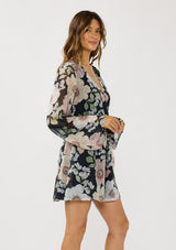 [Color: Black/Blush] A side facing image of a brunette model wearing a black and pink floral mini dress with long bell sleeves, a surplice front, and an elastic waist with side tie detail. A flowy chiffon mini dress for brunch and special occasions. 