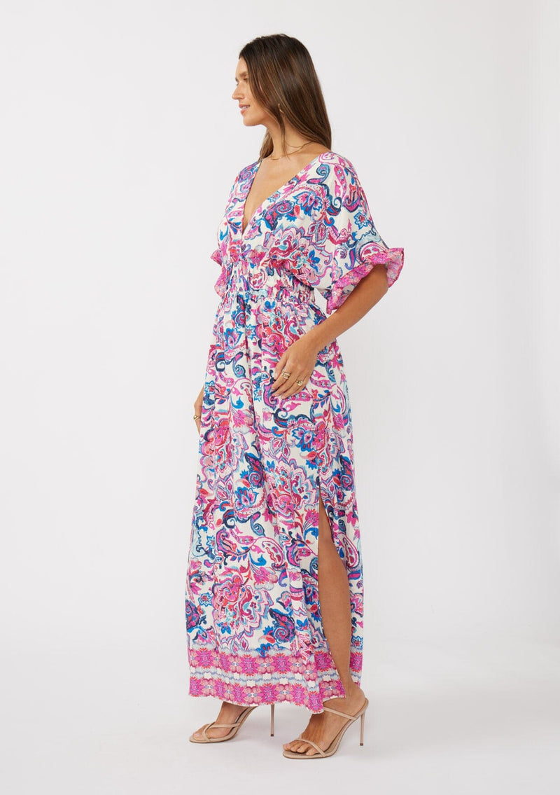 [Color: Natural/Dusty Pink] A side facing image of a brunette model wearing a pink and blue floral print kimono sleeve maxi dress with a ruffle trim, smocked waist, double v-neckline, and side slits. Perfect for spring special occasions. 