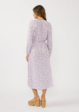 [Color: Natural/Lilac] A back facing image of a brunette model wearing a a purple floral long sleeve midi dress with a button front, a drawstring waist, and side pockets. Flowy and relaxed with side slits for easy movement. 
