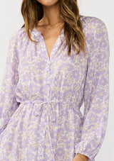[Color: Natural/Lilac] A detail image of a brunette model wearing a a purple floral long sleeve midi dress with a button front, a drawstring waist, and side pockets. Flowy and relaxed with side slits for easy movement. 