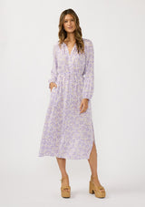 [Color: Natural/Lilac] A front facing image of a brunette model wearing a a purple floral long sleeve midi dress with a button front, a drawstring waist, and side pockets. Flowy and relaxed with side slits for easy movement. 