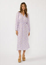 [Color: Natural/Lilac] A front facing image of a brunette model wearing a a purple floral long sleeve midi dress with a button front, a drawstring waist, and side pockets. Flowy and relaxed with side slits for easy movement. 