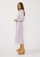 [Color: Natural/Lilac] A side facing image of a brunette model wearing a a purple floral long sleeve midi dress with a button front, a drawstring waist, and side pockets. Flowy and relaxed with side slits for easy movement. 