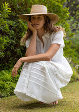 [Color: White] A sitting image of a blonde model in a white cotton midi dress featuring a collared neckline, button front, self-tie waist belt, lace trim, and pintuck details. A timeless boho spring style.