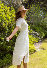 [Color: White] A side facing image of a blonde model in a white cotton midi dress featuring a collared neckline, button front, self-tie waist belt, lace trim, and pintuck details. A timeless boho spring style.