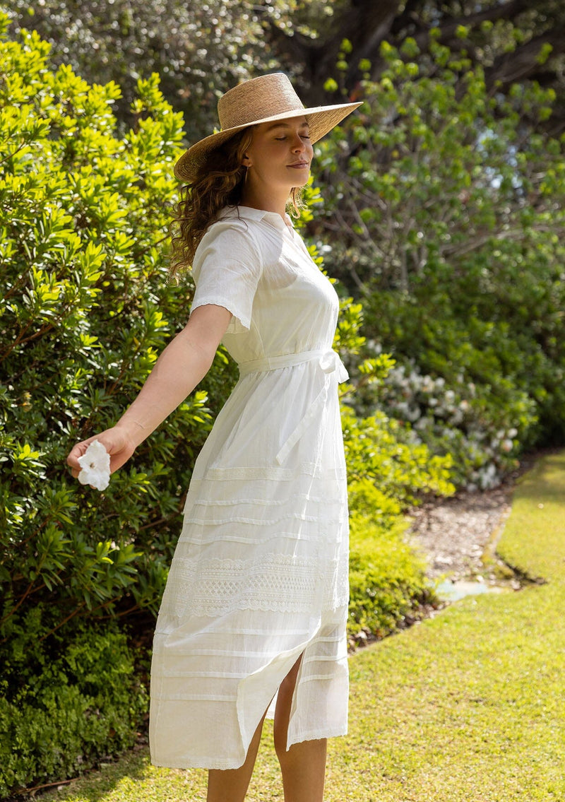 [Color: White] A side facing image of a blonde model in a white cotton midi dress featuring a collared neckline, button front, self-tie waist belt, lace trim, and pintuck details. A timeless boho spring style.