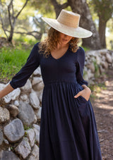 [Color: Navy] A blonde model wearing a navy maxi dress with a smocked bodice, v-neckline, 3/4 length sleeves, and side pockets. The perfect cotton maxi dress for any season.