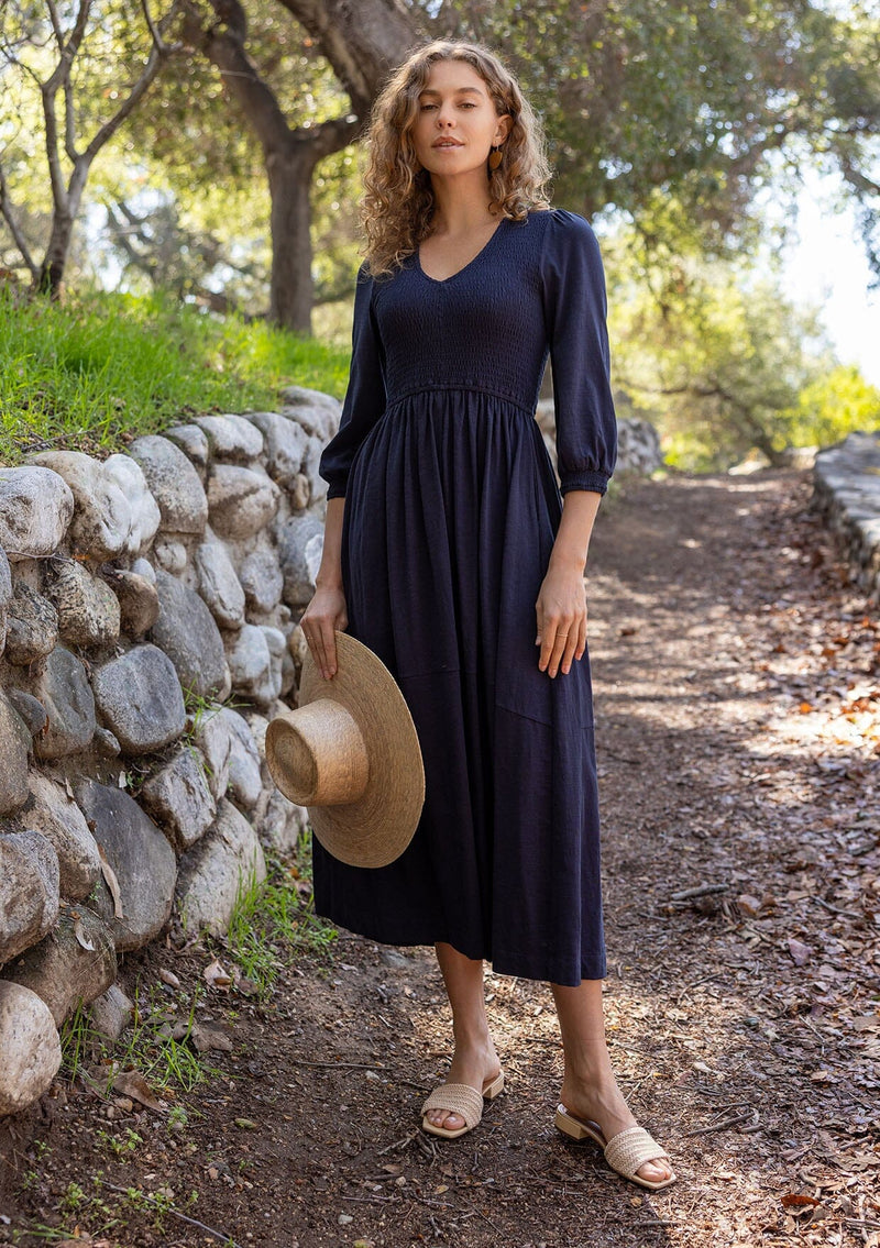 [Color: Navy] A blonde model wearing a navy maxi dress with a smocked bodice, v-neckline, 3/4 length sleeves, and side pockets. The perfect cotton maxi dress for any season.