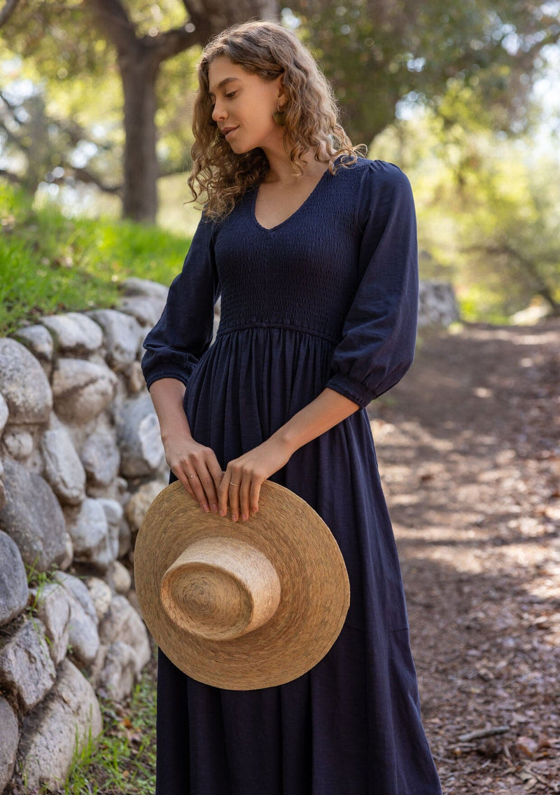 [Color: Navy] A blonde model wearing a navy maxi dress with a smocked bodice, v-neckline, 3/4 length sleeves, and side pockets. The perfect cotton maxi dress for any season.