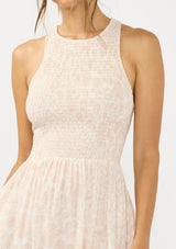 [Color Shell/Cream] A detail image of a brunette model wearing a floral smocked midi dress with a sleeveless design, round neck, side pockets, and a cross back with tie detail. 
