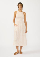 [Color Shell/Cream] A front facing image of a brunette model wearing a floral smocked midi dress with a sleeveless design, round neck, side pockets, and a cross back with tie detail. 