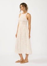 [Color Shell/Cream] A side facing image of a brunette model wearing a floral smocked midi dress with a sleeveless design, round neck, side pockets, and a cross back with tie detail. 