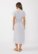 [Color: Dove] A back facing image of a brunette model wearing a light purple midi dress with short flutter sleeves, a v-neckline, a decorative button front, and a smocked waistline. 