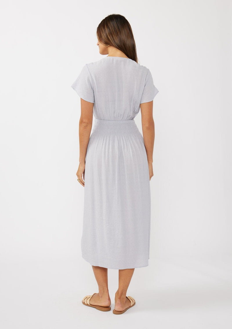 [Color: Dove] A back facing image of a brunette model wearing a light purple midi dress with short flutter sleeves, a v-neckline, a decorative button front, and a smocked waistline. 