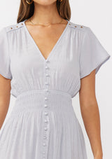 [Color: Dove] A detail image of a brunette model wearing a light purple midi dress with short flutter sleeves, a v-neckline, a decorative button front, and a smocked waistline. 