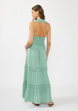 [Color: Navy] A back facing image of a brunette model wearing a flowy green maxi dress with a halter neck, v-neckline, ruffle details, and a tiered skirt. 