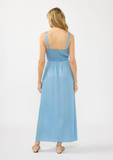 [Color: Light Wash] A back facing image of a blonde model wearing a blue tencel maxi dress with a sleeveless design, snap button front, ruched bodice, and a smocked back. 