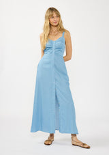 [Color: Light Wash] A front facing image of a blonde model wearing a blue tencel maxi dress with a sleeveless design, snap button front, ruched bodice, and a smocked back. 