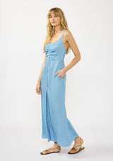 [Color: Light Wash] A side facing image of a blonde model wearing a blue tencel maxi dress with a sleeveless design, snap button front, ruched bodice, and a smocked back. 