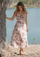 [Color: Almond/Wine] A front facing image of a blonde model by a lake wearing a pink floral print midi dress with a scooped neckline, smocked bust and back, elastic shoulder straps with ruffle trim, and a sexy cutout back detail. 