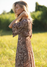 [Color: Natural/Brown] A side facing image of a blonde model wearing a brown paisley maxi dress with a deep v-neckline, flowy short sleeves, twist front, and a side slit. A fall bohemian maxi dress for special occasions. 