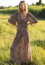[Color: Natural/Brown] A front facing image of a blonde model wearing a brown paisley maxi dress with a deep v-neckline, flowy short sleeves, twist front, and a side slit. A fall bohemian maxi dress for special occasions. 