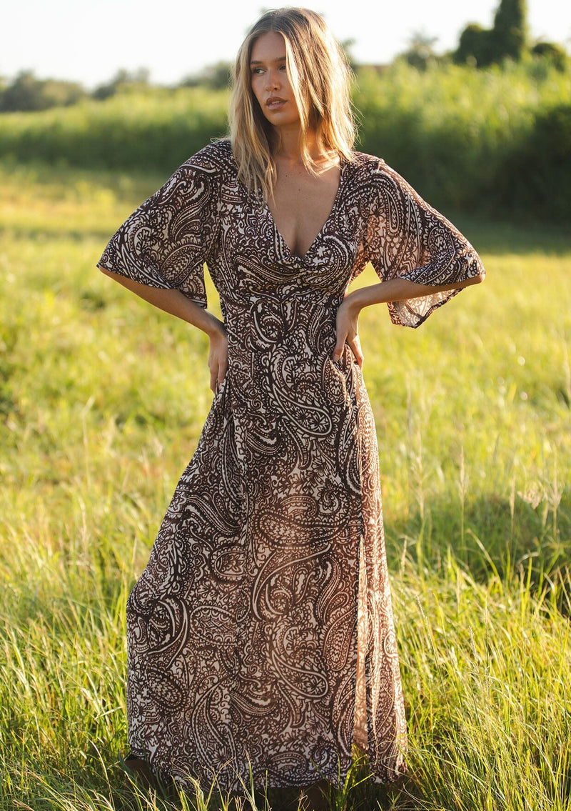[Color: Natural/Brown] A front facing image of a blonde model wearing a brown paisley maxi dress with a deep v-neckline, flowy short sleeves, twist front, and a side slit. A fall bohemian maxi dress for special occasions. 