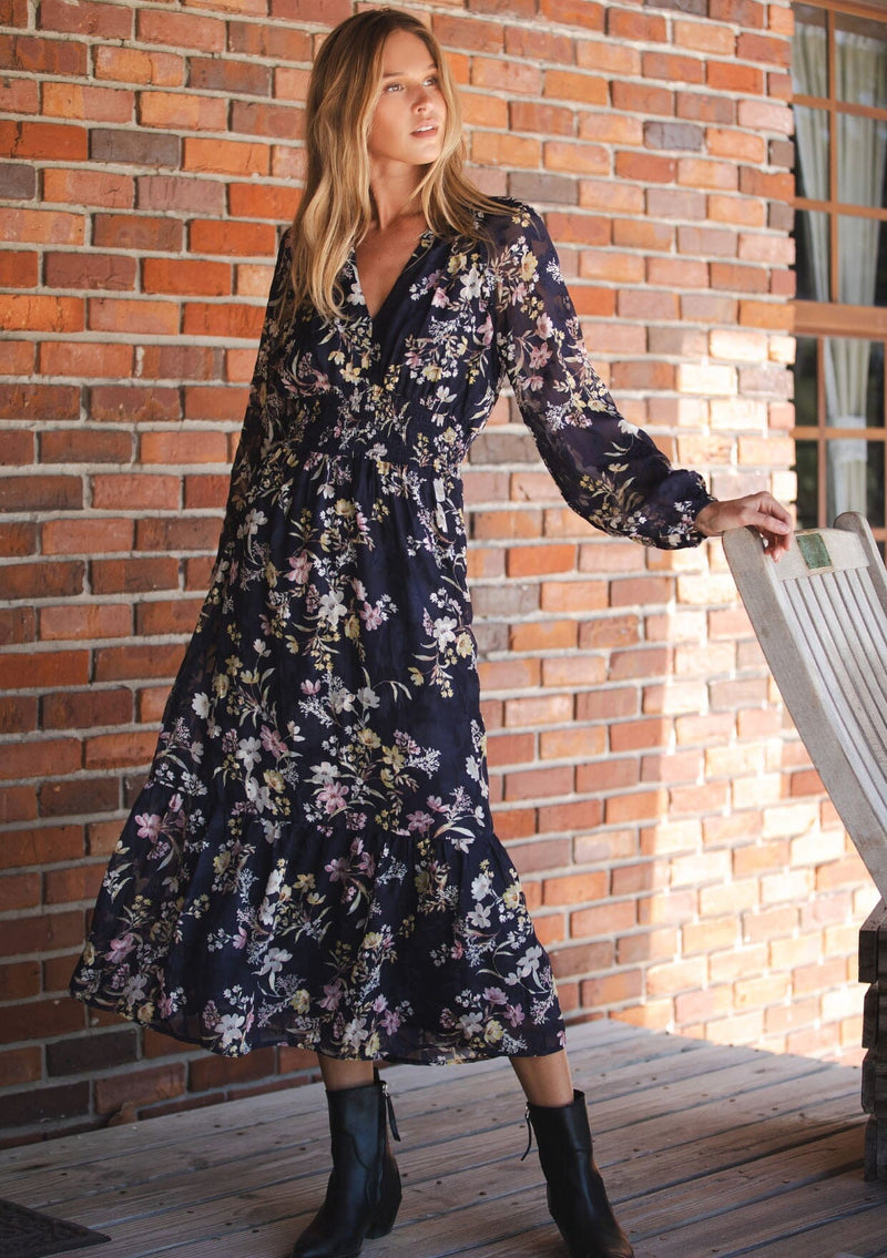 [Color: Navy/Yellow] A front facing image of a blonde model wearing a navy floral maxi dress with long sleeves, a smocked waist, and a split v-neckline. A classic floral maxi for weddings and special occasions. 