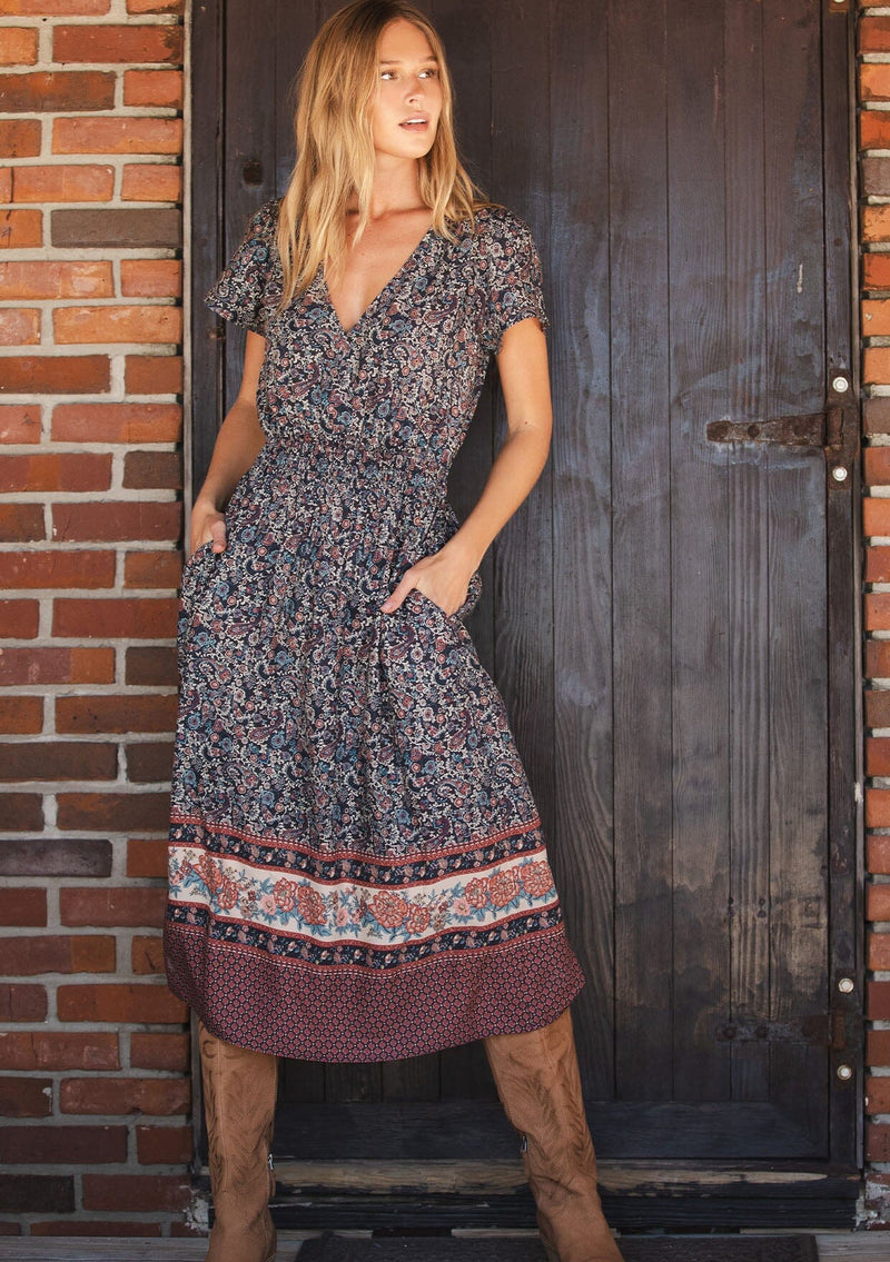 [Color: Navy/Rose] A front facing image of a blonde model wearing a navy floral border print midi dress with a classic v-neckline, short sleeves, a smocked waist, and side pockets. A fall bohemian dress perfect for any occasion. 