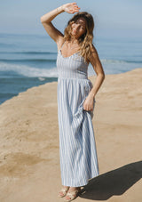 [Color: Blue/White] A side facing image of a blonde model at the beach wearing blue and white striped midi dress with a smocked bodice, v-neckline, and adjustable straps. A breezy cotton blend dress for vacation. 