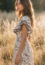 [Color: Natural/Olive] A side facing image of a brunette model in the fields wearing a beige and black floral print midi dress with short flutter sleeves, a v neckline, and a ruffle trimmed hem. 