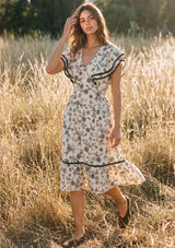 [Color: Natural/Olive] A front facing image of a brunette model in the fields wearing a beige and black floral print midi dress with short flutter sleeves, a v neckline, and a ruffle trimmed hem. 