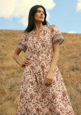 [Color: Natural/Brown] A front facing image of a brunette model wearing a brown bohemian floral border print midi dress with a split v-neckline, short sleeves, and an elastic waist. A fall boho dress styled with boots. 