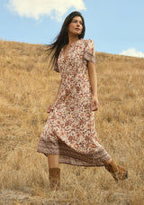 [Color: Natural/Brown] A side facing image of a brunette model wearing a brown bohemian floral border print midi dress with a split v-neckline, short sleeves, and an elastic waist. A fall boho dress styled with boots. 