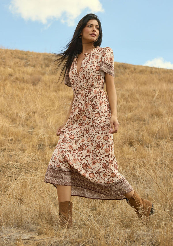 [Color: Natural/Brown] A side facing image of a brunette model wearing a brown bohemian floral border print midi dress with a split v-neckline, short sleeves, and an elastic waist. A fall boho dress styled with boots. 