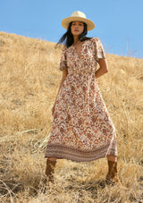 [Color: Natural/Brown] A front facing image of a brunette model wearing a brown bohemian floral border print midi dress with a split v-neckline, short sleeves, and an elastic waist. A fall boho dress styled with boots. 
