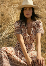 [Color: Natural/Brown] A detail sitting image of a brunette model wearing a brown bohemian floral border print midi dress with a split v-neckline, short sleeves, and an elastic waist. A fall boho dress styled with boots. 
