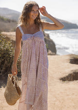 [Color: Vanilla/Rose] A front facing image of a blonde model at the beach wearing a pink floral bohemian maxi dress with a flowy tiered silhouette, crochet adjustable straps, a ruched bust detail, and ruffle trim accents. A pretty beach dress for vacation. 