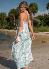 [Color: Natural/Mint] A back facing image of a blonde model at the beach wearing a sleeveless mint green floral maxi dress with a tiered silhouette, high low hemline, ruffle trims, and side pockets. 