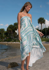 [Color: Natural/Mint] A front facing image of a blonde model at the beach wearing a sleeveless mint green floral maxi dress with a tiered silhouette, high low hemline, ruffle trims, and side pockets. 