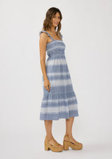 [Color: Navy/White] A side facing image of a brunette model wearing a blue and white checkered midi dress with stripes. Featuring chic ruffled elastic straps, a smocked bust, side pockets, and a tiered skirt. 