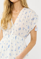 [Color: White/Blue] A detail image of a blonde model wearing a spring white and blue floral maxi dress with short dolman sleeves, tassel ties, a v neckline, and a smocked waist. 