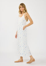[Color: White/Blue] A side facing image of a blonde model wearing a white and blue floral maxi dress with a relaxed fit, scoop neckline, elastic shoulder straps, and  side pockets.