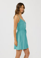 [Color: Teal] A side facing image of a brunette model wearing a teal sleeveless mini dress with spaghetti straps, a cross back, button front, drawstring waist, and side pockets. 