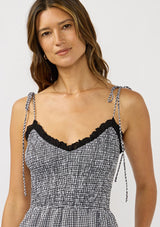[Color: Black/White] A  detail image of a brunette model wearing a black and white gingham midi dress with a smocked bodice, tie shoulder straps, and side pockets. 